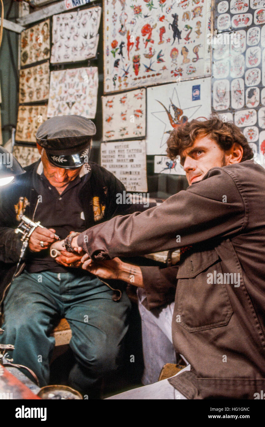 A customer shows a brave expression as he gets a painful tattoo at a state fair in Rochester, New Hampshire. Note exhibition of tattoo designs on wall. Stock Photo