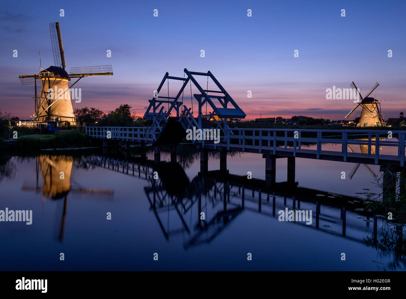 Two illuminated windmills and a drawbridge with reflection in the water of the canal in the last light at sunset in Kinderdijk in the Netherlands. Stock Photo