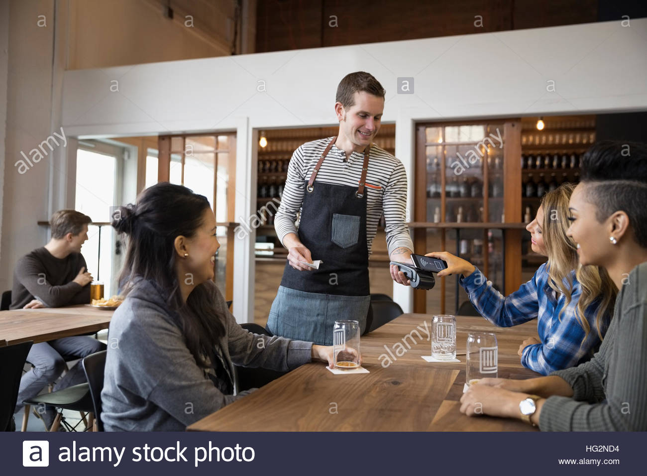Female friends paying waiter with smart phone contactless payment in brewery tasting room restaurant Stock Photo