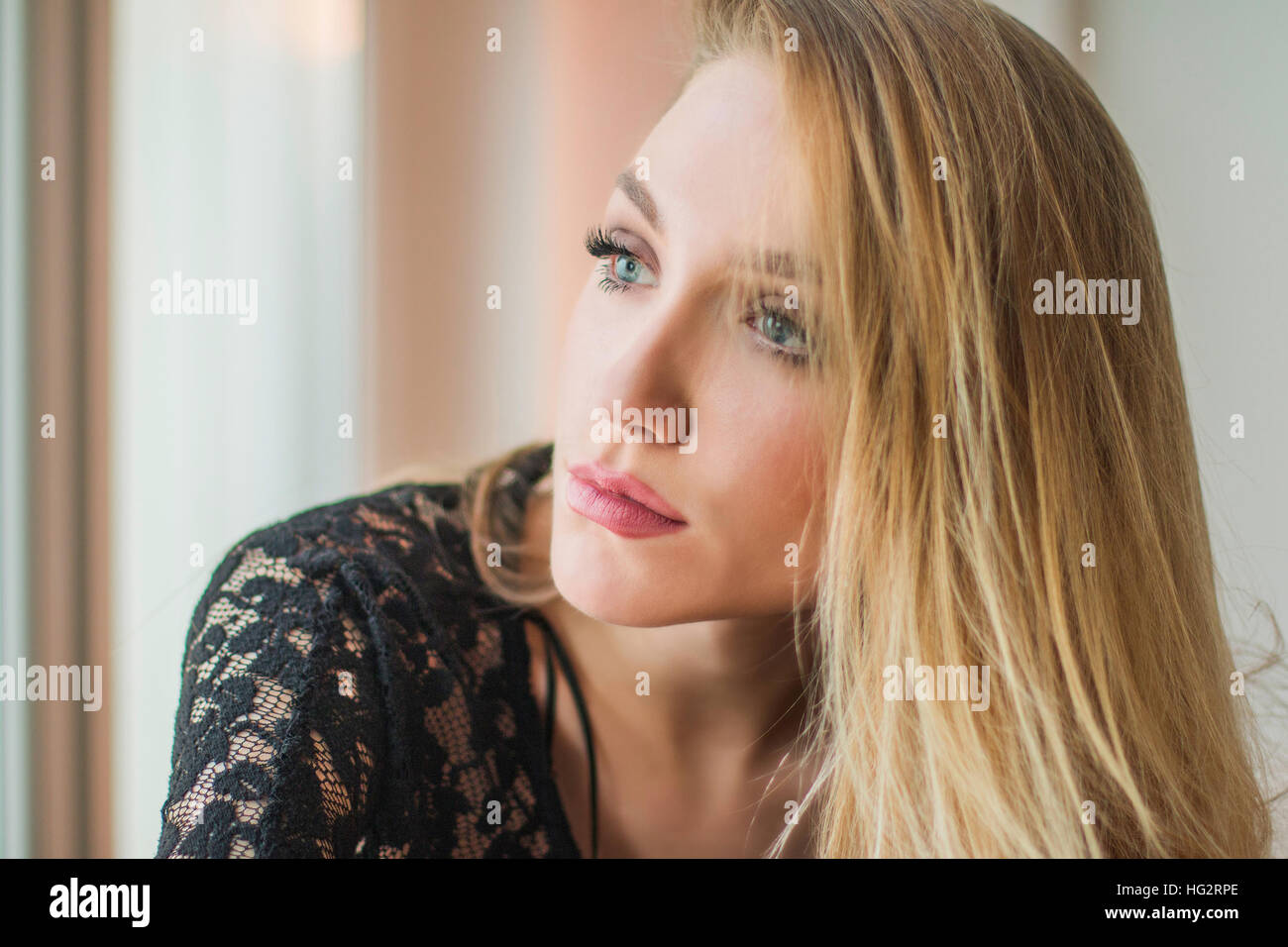 Close up portrait of beautiful young blonde woman in black lace shirt sitting next to window, looking emotive and thoughtful Stock Photo