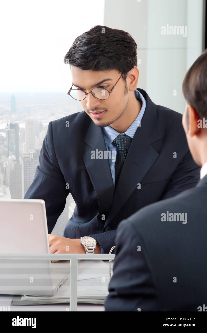 Young professional man working on laptop computer in office cabin while another professional man is sitting opposite to him Stock Photo