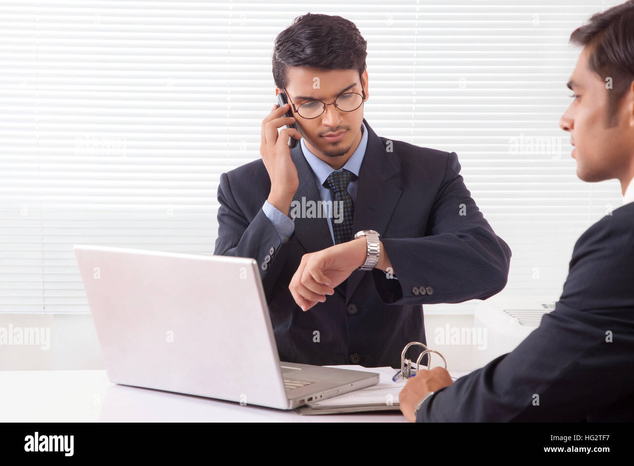 Young professional man looking at wrist watch while talking over mobile phone at office while another professional man is sitting opposite to him Stock Photo