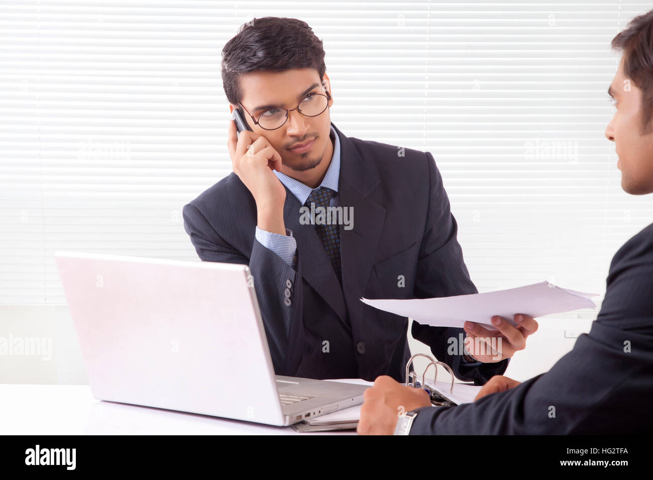 Young professional man handing over document to another professional man while talking over mobile phone in office Stock Photo