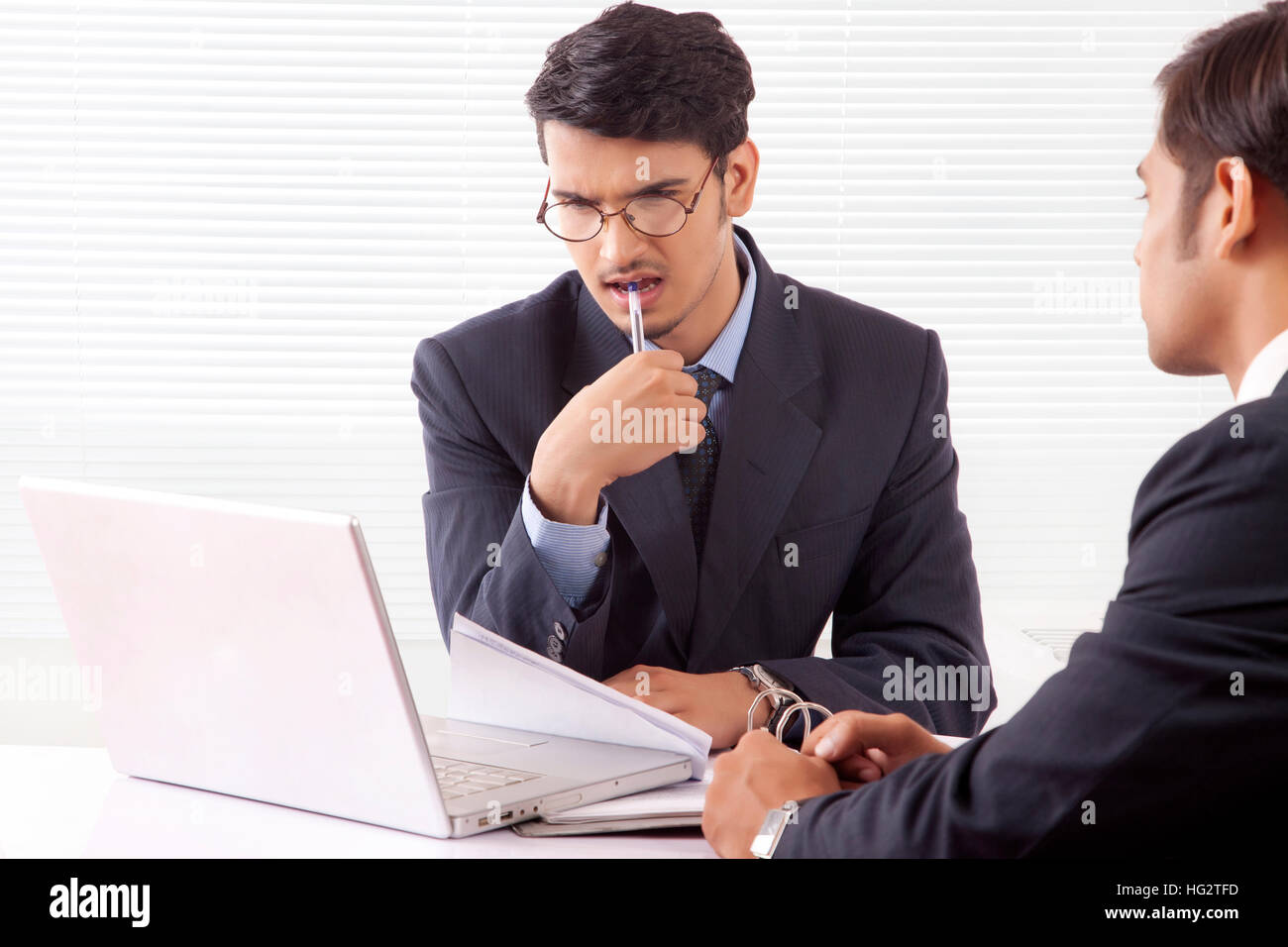 Young professional man in deep thought holding pen between his teeth looking at laptop computer while discussing work with another professional man Stock Photo
