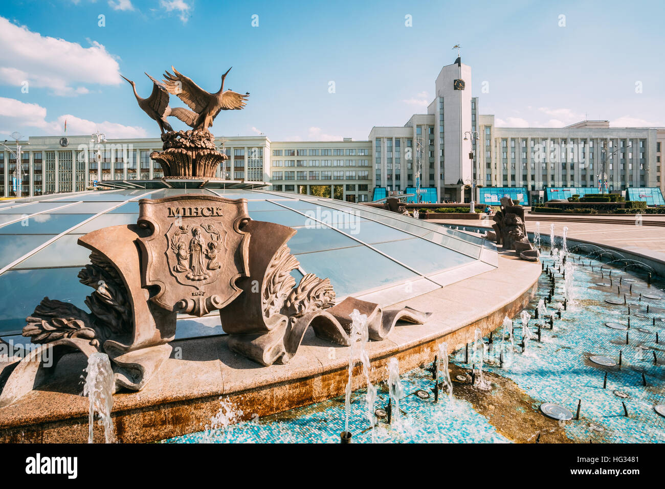 Minsk, Belarus. Fountains Around Glass Dome Of Stolitsa Underground Shopping Center At Independence Square With Bronze Coat Of Arms Of Minsk In Summer Stock Photo