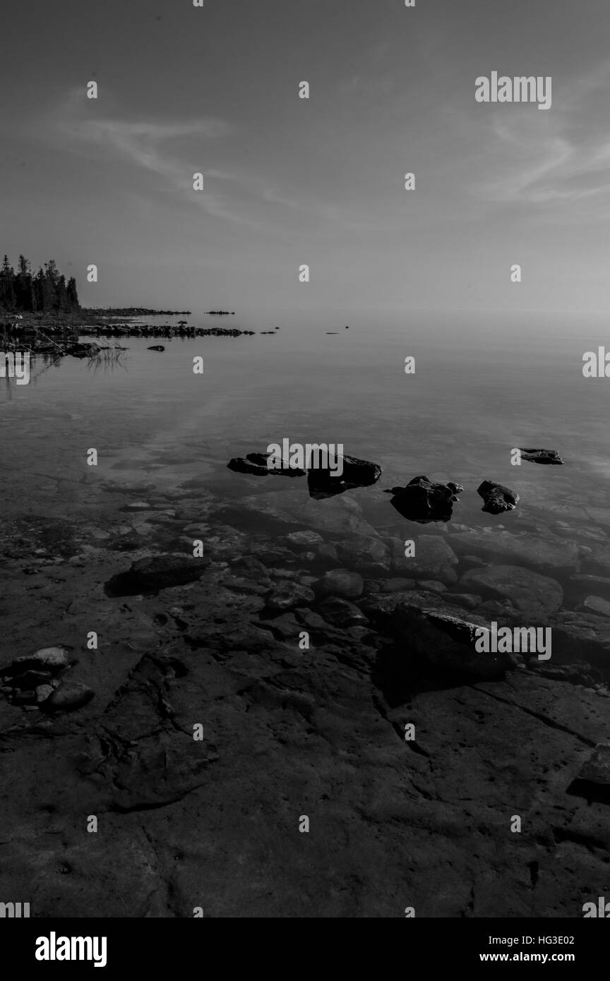 Dead calm afternoon image of clear lake huron water and limestone rocks along shoreline.  Clouds reflecting in a zign zag. b/w Stock Photo