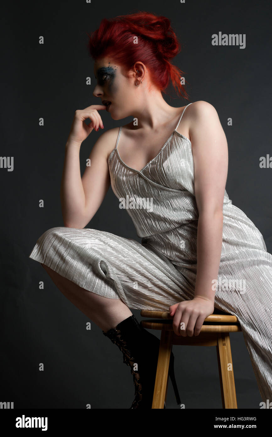 Red haired model with blue creative makeup wearing silver jumpsuit, seated on wooden stool Stock Photo