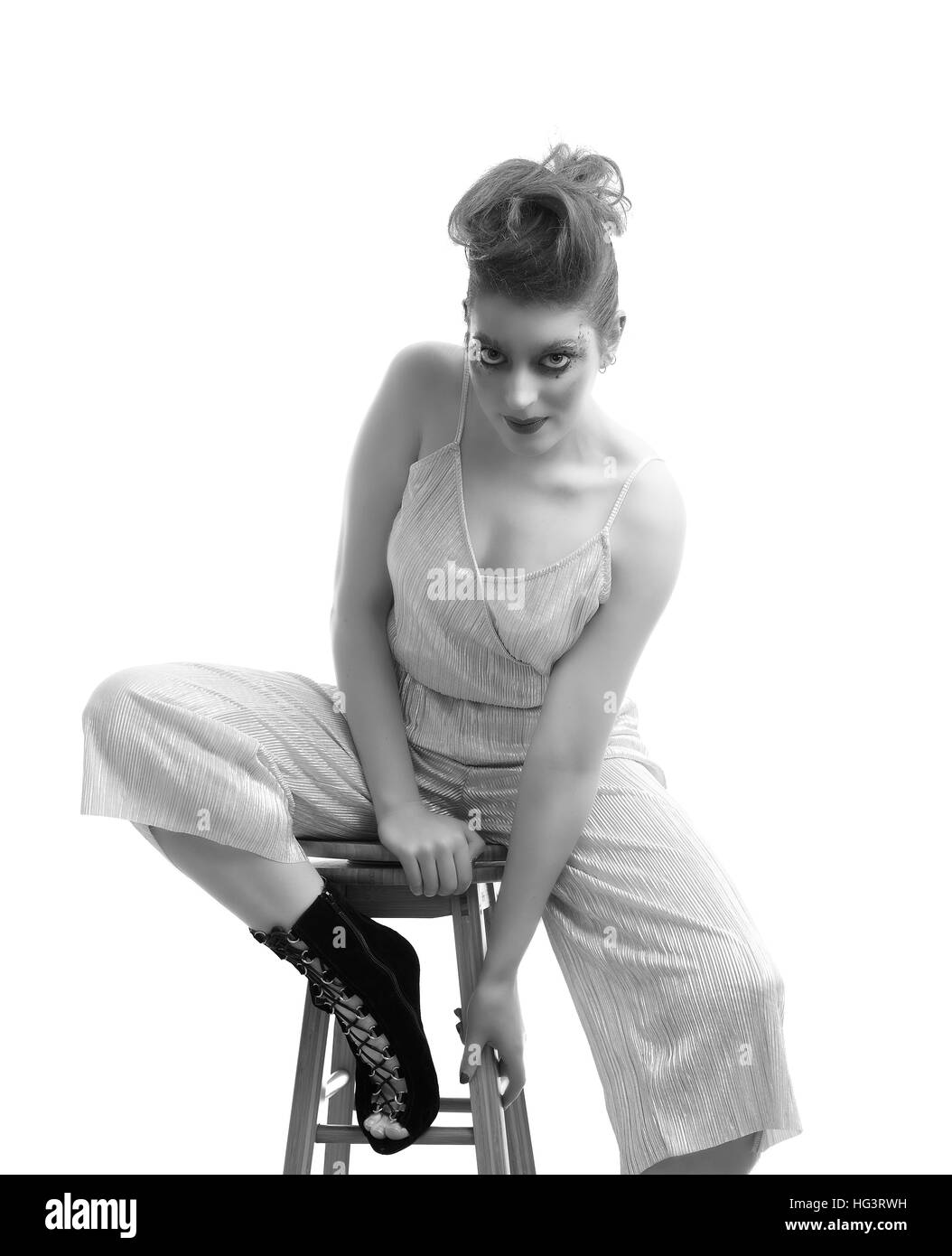 Model with creative makeup wearing silver jumpsuit, seated on wooden stool, monochrome Stock Photo