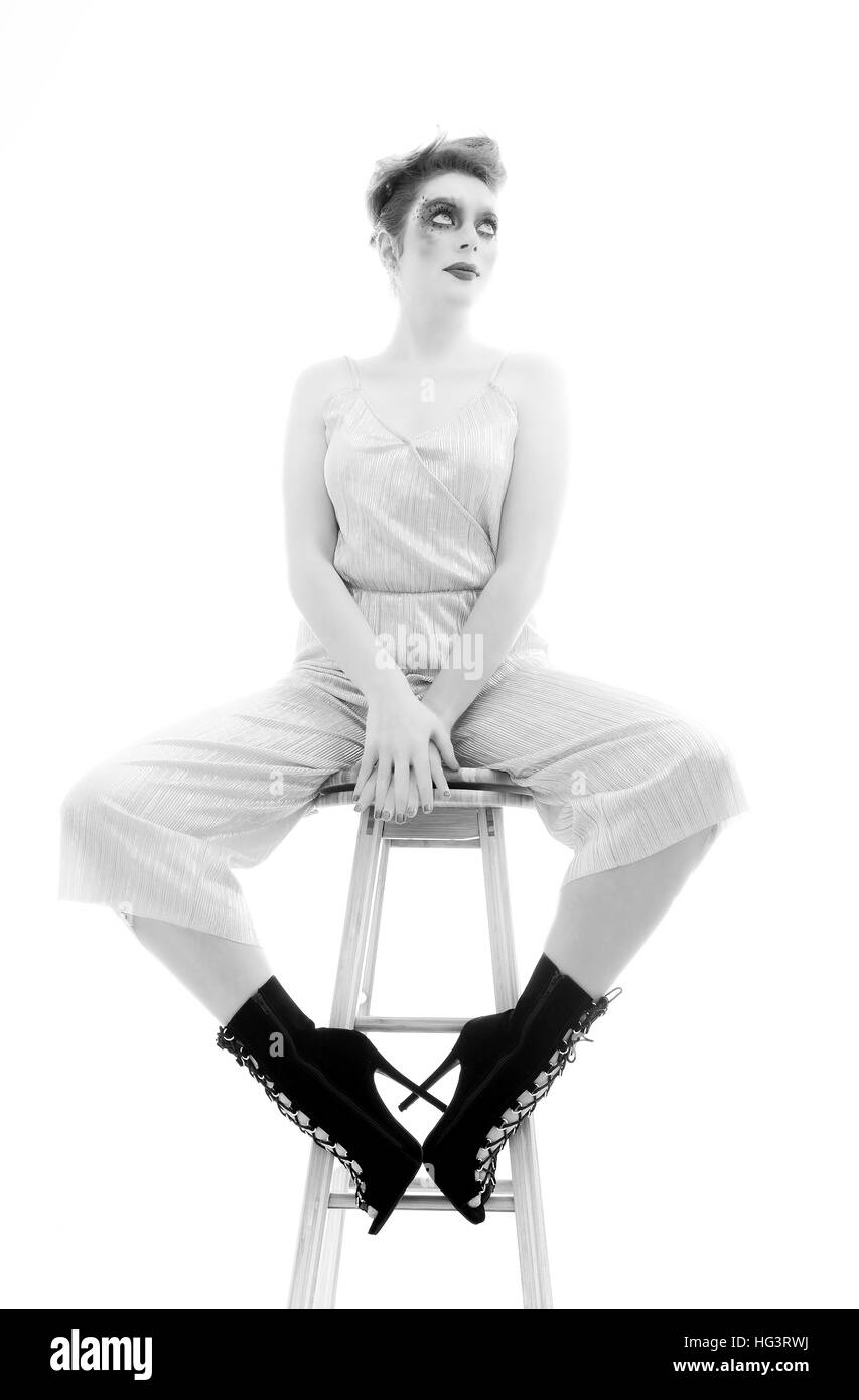 Model with creative makeup wearing silver jumpsuit, seated on wooden stool, monochrome full length Stock Photo