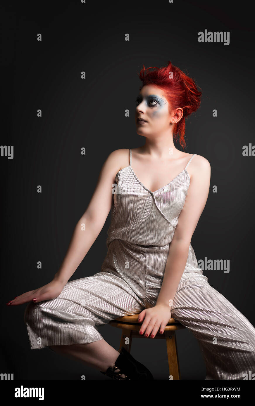 Red haired model with blue creative makeup wearing silver jumpsuit, seated on wooden stool Stock Photo