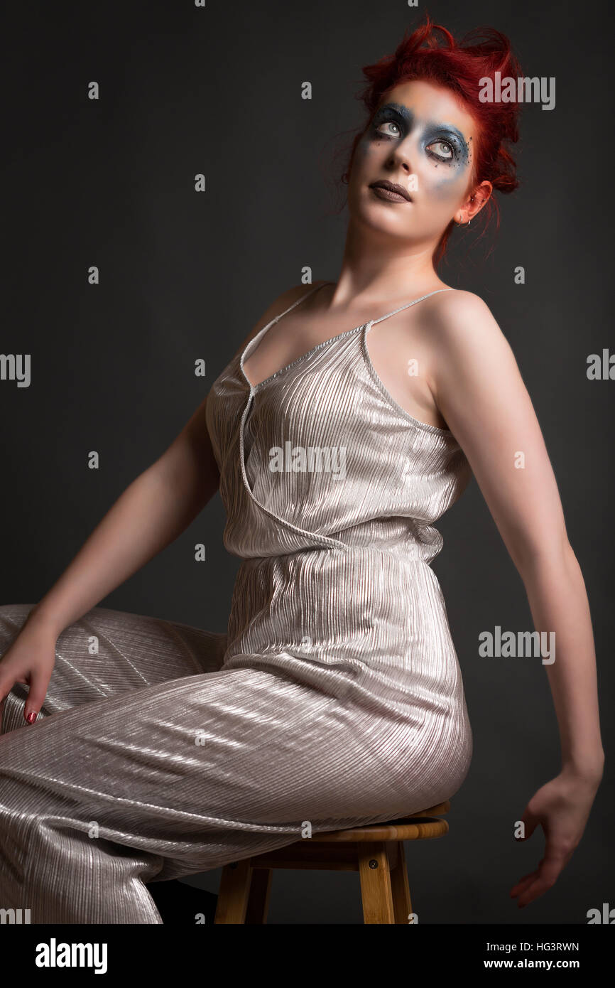 Red haired model with blue creative makeup wearing silver jumpsuit, seated on wooden stool Stock Photo