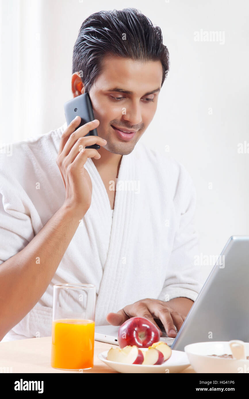 Man talking on phone and working on laptop at breakfast table Stock Photo