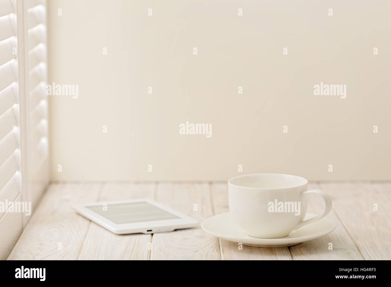 E-book and a cup on a light wooden background near a window with shutters. Stock Photo