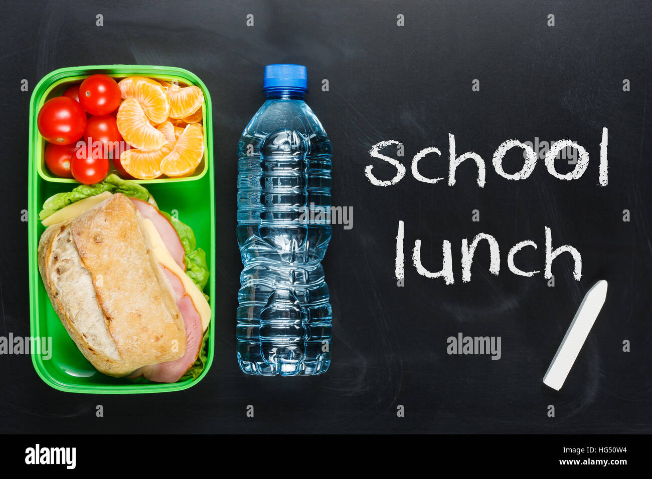 Sandwich, small tomatoes, tangerine in plastic lunch box and bottle of water on black chalkboard. Stock Photo