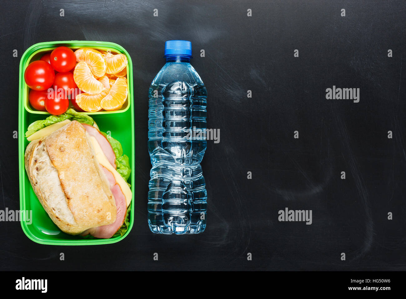 Sandwich, small tomatoes, tangerine in plastic lunch box and bottle of water on black chalkboard. Stock Photo