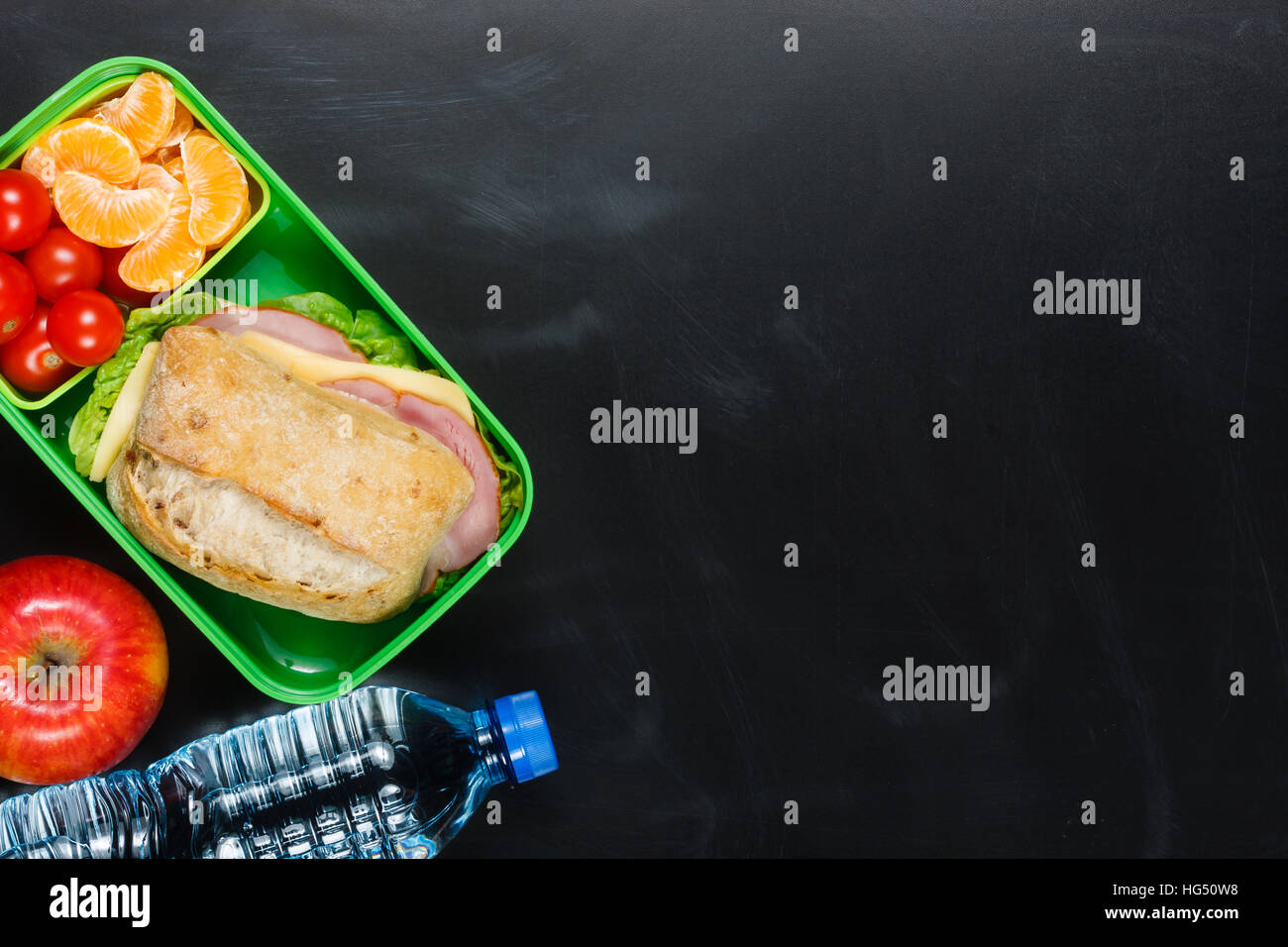 Sandwich, small tomatoes, tangerine, apple in plastic lunch box and bottle of water on black chalkboard. Stock Photo