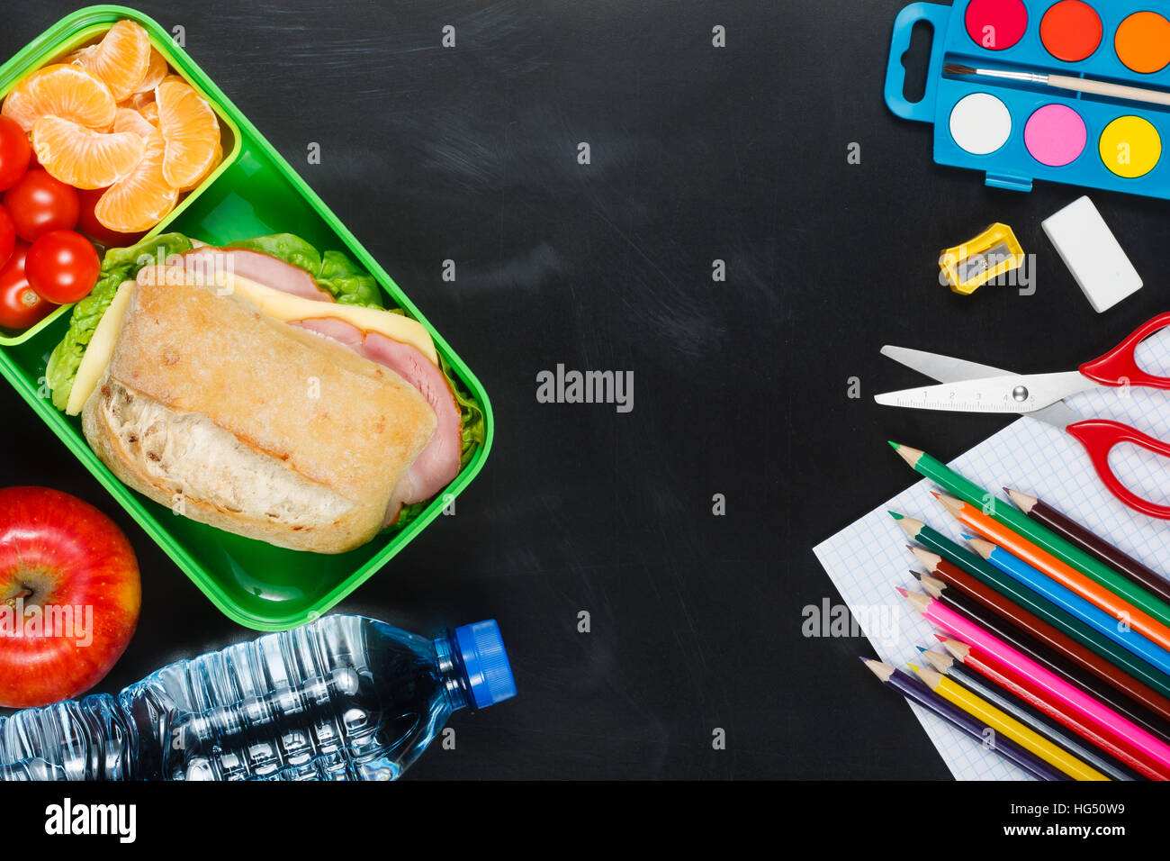 School lunch. Sandwich, small tomatoes, tangerine, apple in plastic lunch box and bottle of water on black chalkboard. Stock Photo