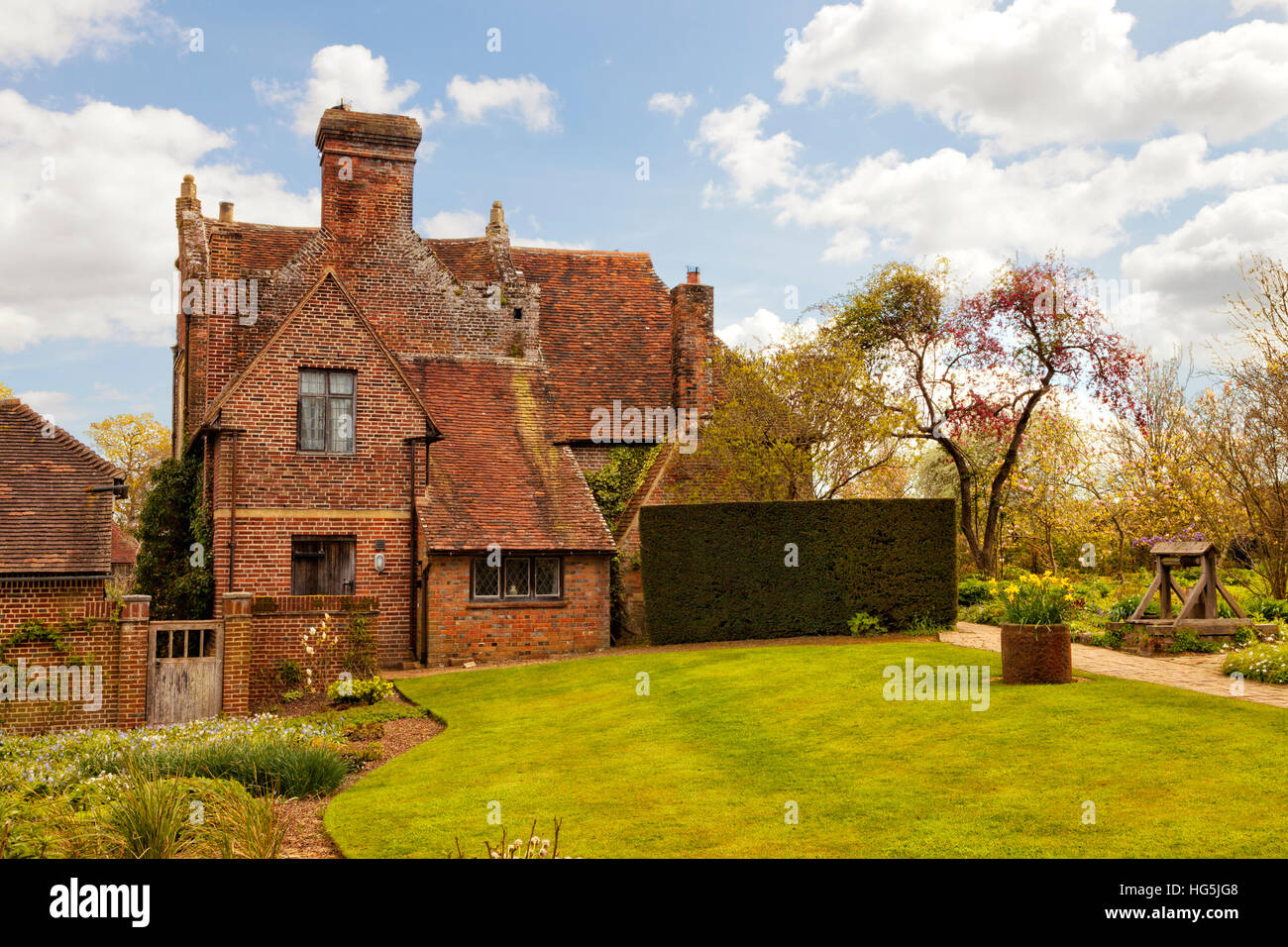 Old red brick english cottage in front of green lawn, with spring garden, water well Stock Photo