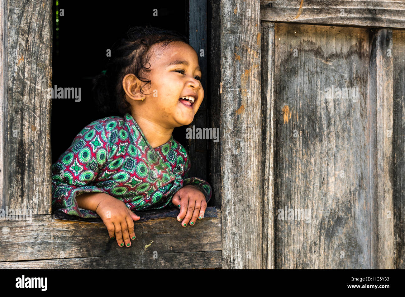 Portrait of loughing little girl, Upper Marsyangdi valley, looking out of a window, Bagarchap, Manang District, Nepal Stock Photo
