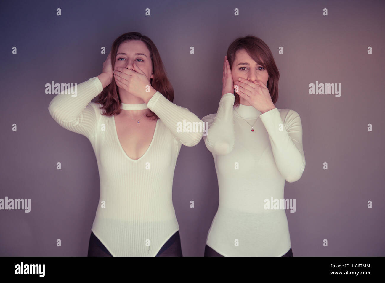 Doppelganger / Alter-ego themed photoshoot: two young women girls wearing  matching clothes stood standing together matching echoing mirroring each  others physical pose and expression - one hand over the mouth and one