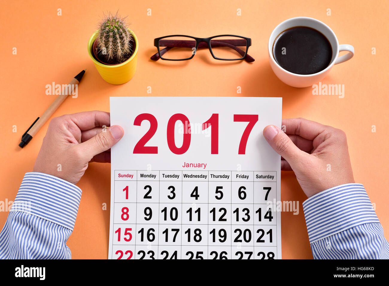 a young caucasian businessman observes a 2017 calendar sitting at his office desk, where there is a pen, a cactus, a pair of eyeglasses and a cup of c Stock Photo