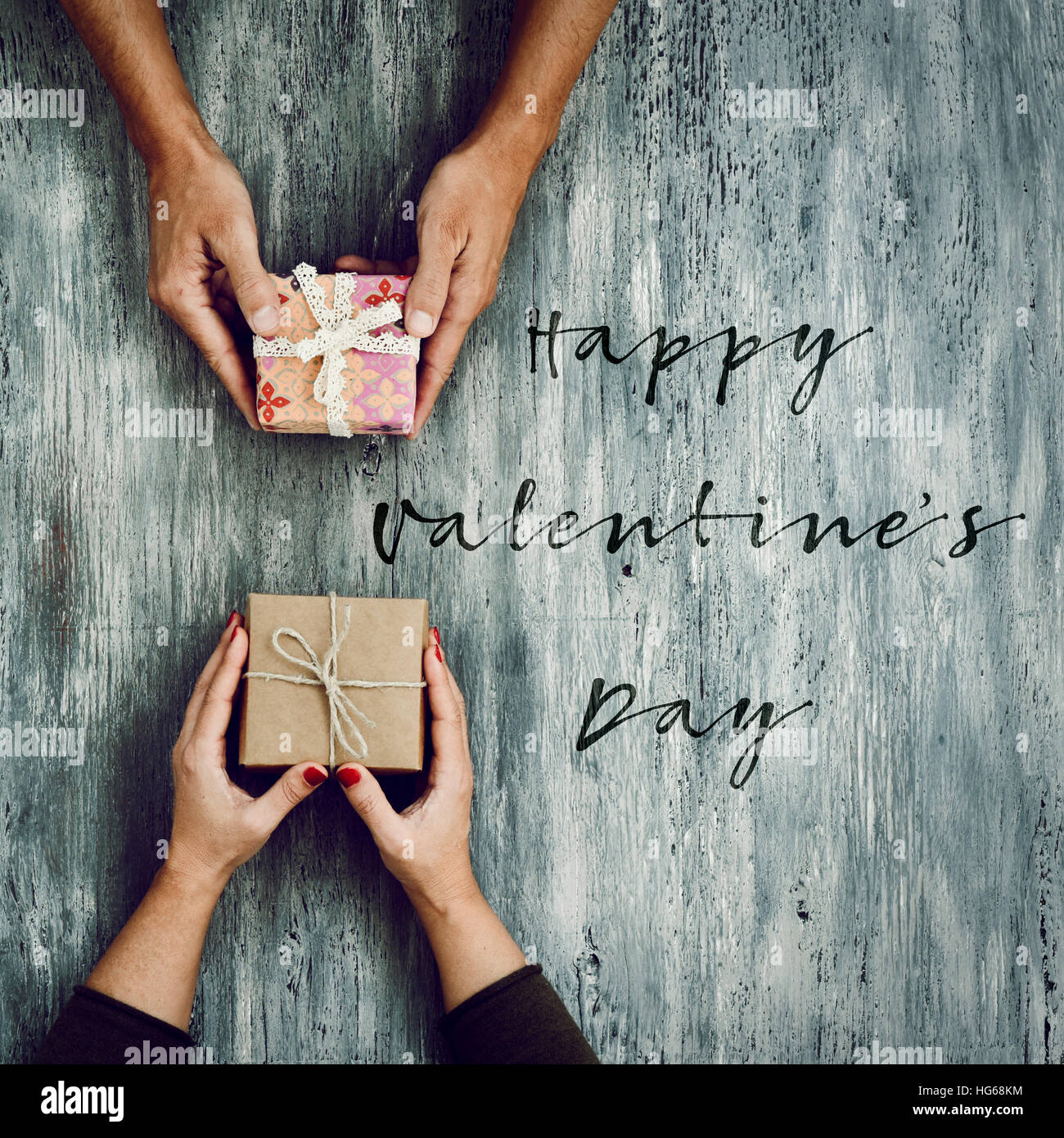 high-angle shot of a young caucasian woman and a young caucasian man exchanging gifts and the text happy valentines day on a rustic wooden table Stock Photo