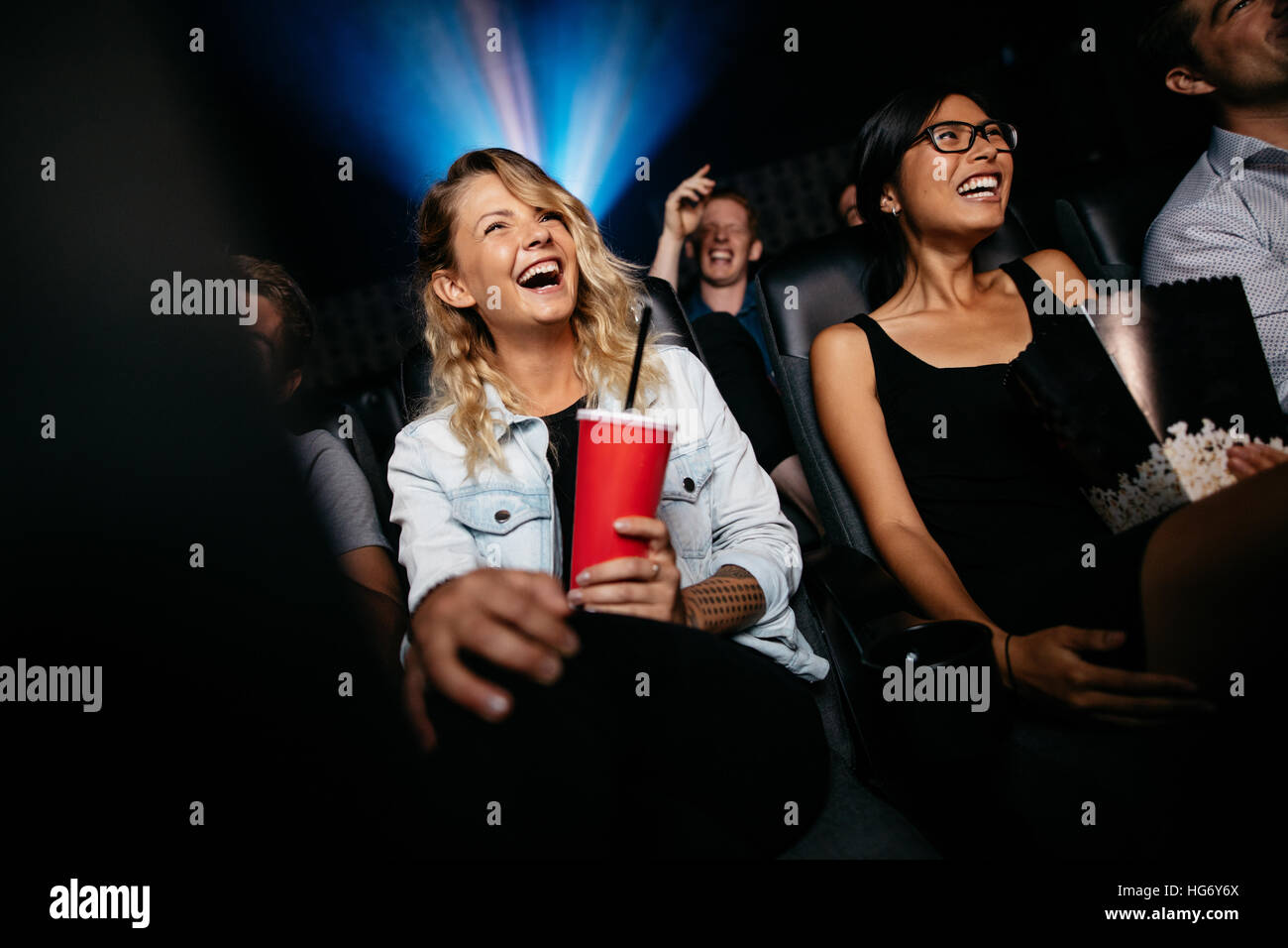 Young people laughing while watching film in movie theater. Group of friends in multiplex cinema with drinking and popcorn. Stock Photo