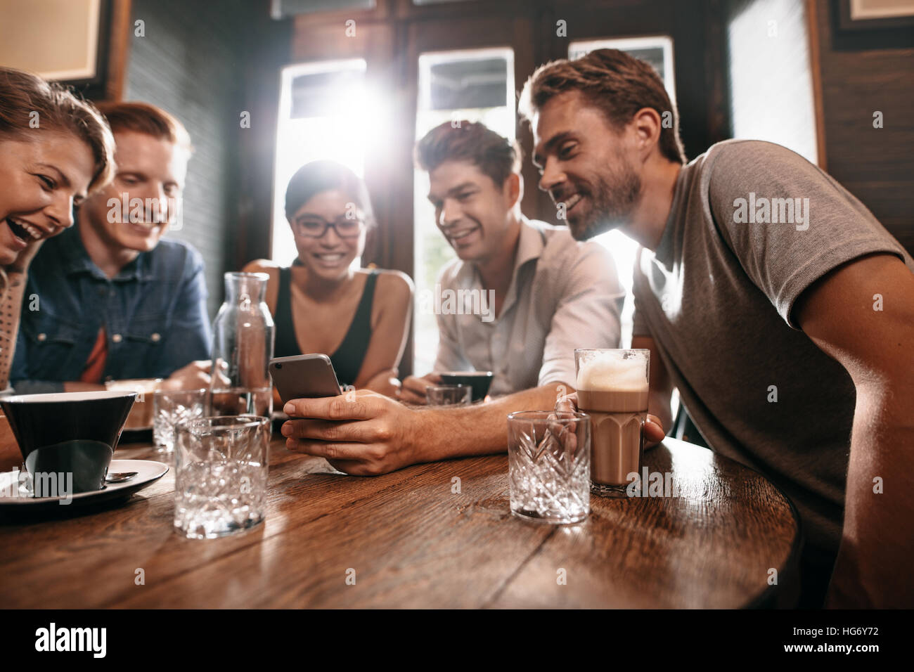 Group of friends sitting around a cafe table and looking at mobile phone. Young men and women looking at pictures on smart phone. Stock Photo