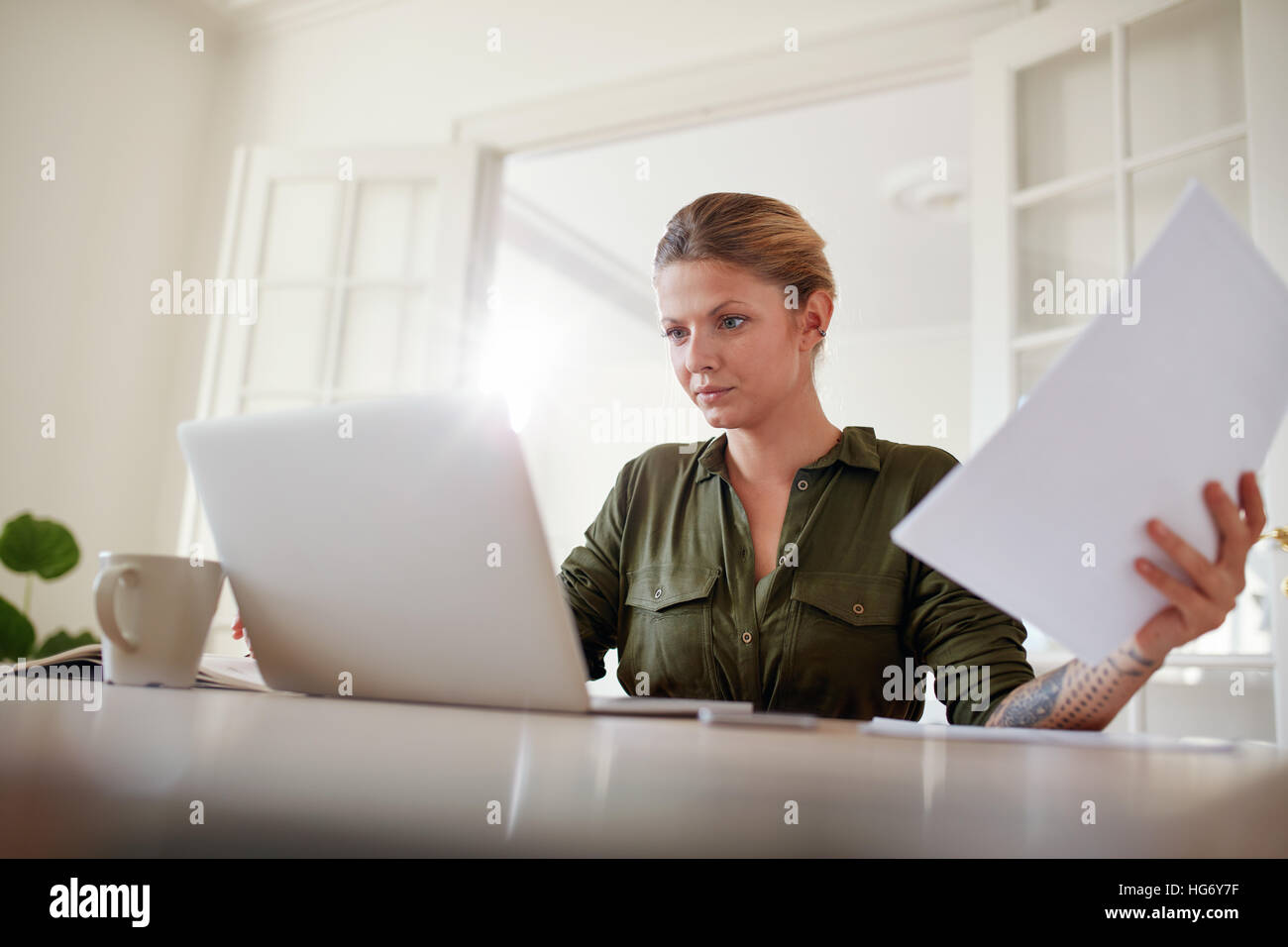 Portrait of young female sitting at table holding documents and working on laptop. Woman busy working at home office. Stock Photo