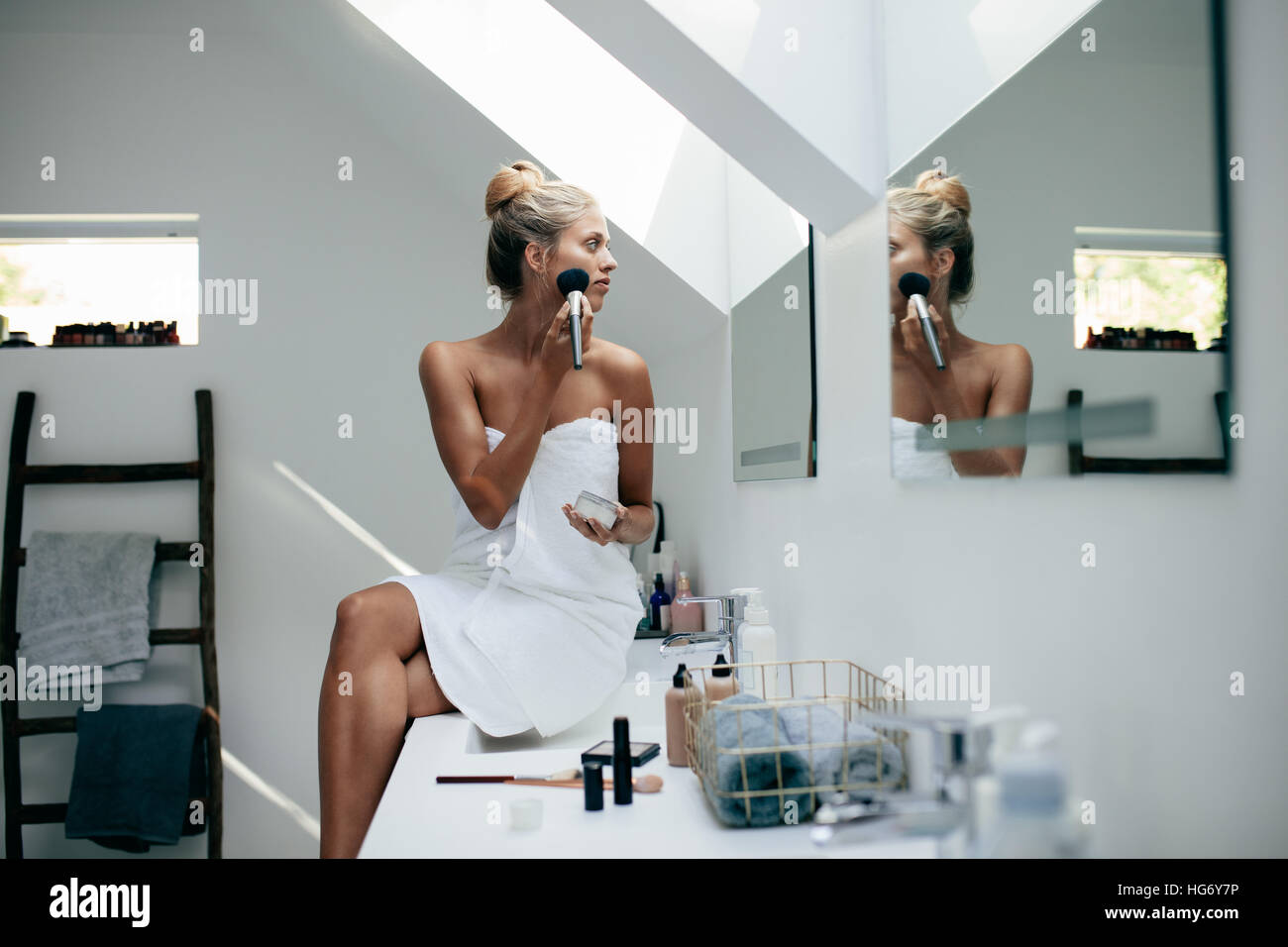 Young woman wrapped in towel with makeup brush looking to mirror at home bathroom and putting on makeup. Stock Photo