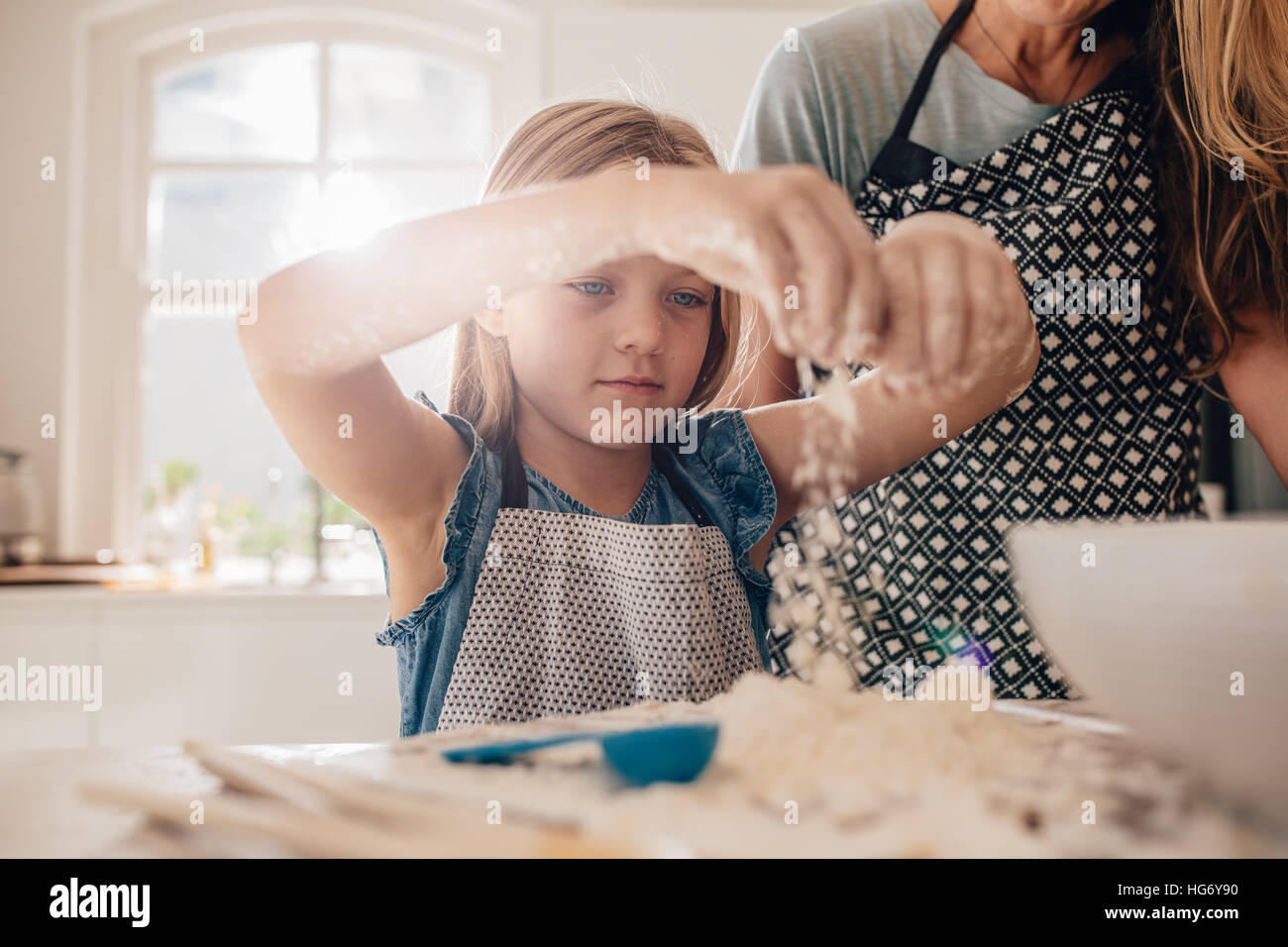 Beautiful little girl learns to cook a meal in the kitchen. Girl making a dough for baking with her mother standing by. Stock Photo