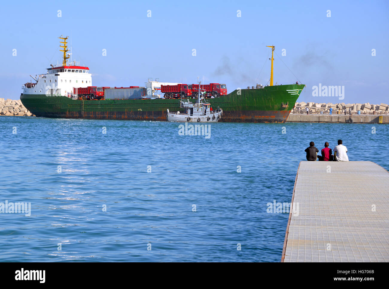 Three boys sitting on a pier watching a small container ship unload cargo, Puntland Somalia Stock Photo