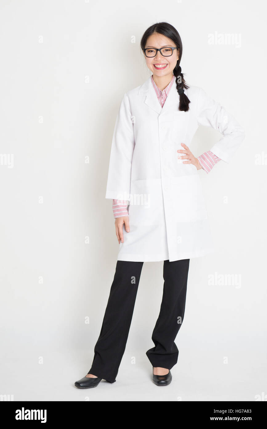 Full length portrait of young Asian female scientist with lab coat standing on plain background. Stock Photo