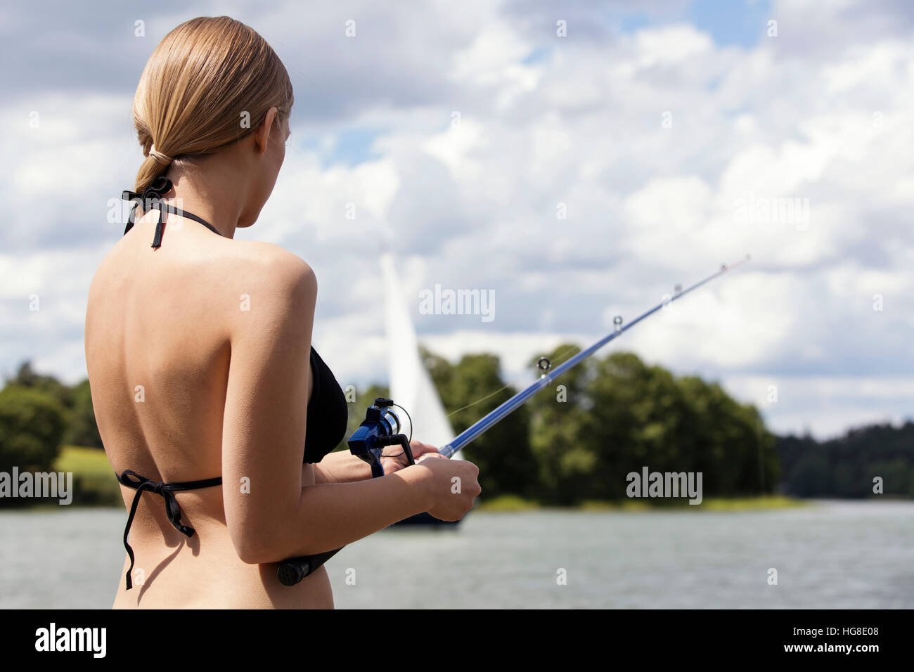 Rear view of woman in bikini fishing while standing against cloudy sky at lakeshore Stock Photo