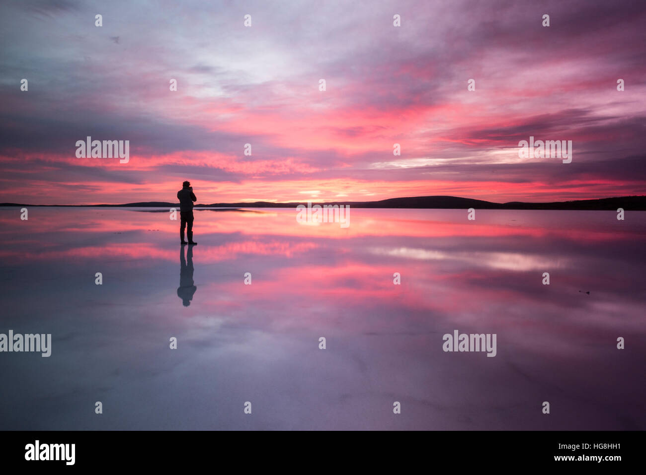 A person stands on a mirror reflected lake watching the sunset Stock Photo