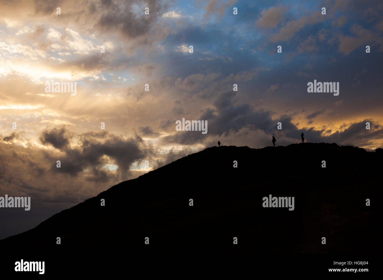 Dramatic silhouette of 3 mountain climbers on the top of a mountain with dramatic cloudy sunset sky. Stock Photo