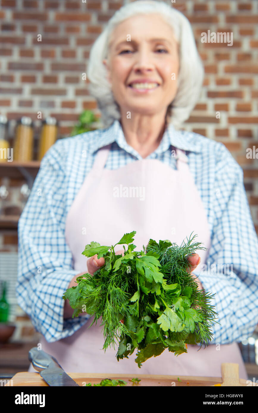 Smiling senior woman holding green herbs in kitchen Stock Photo