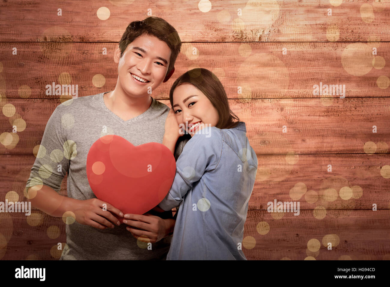 Smiling asian couple in love holding red heart symbol against wooden ...