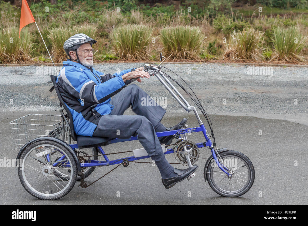 A smiling, elderly man, age 94, pedals a recumbent tricycle along a driveway, equipped with a helmet, flag and other safety gear. Stock Photo