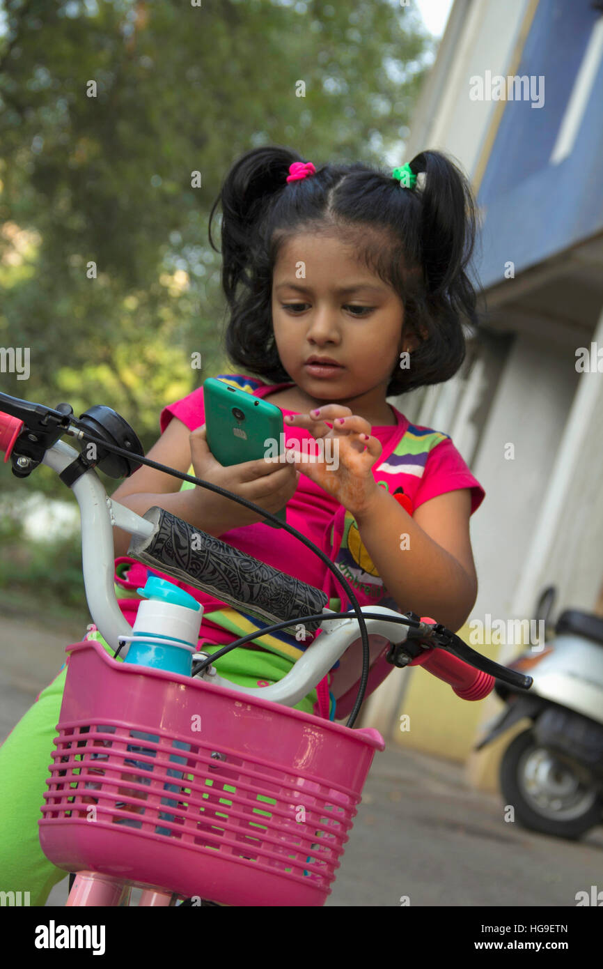 Little Indian girl checking mobile while sitting on bicycle Stock Photo