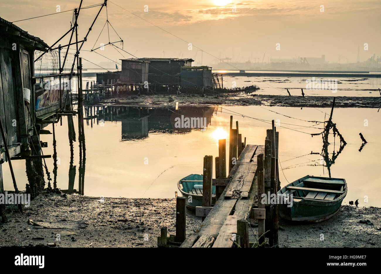 Old fishing huts reflecting in the lagoon of Marina di Ravenna at sunset, industrial harbor on background. Stock Photo