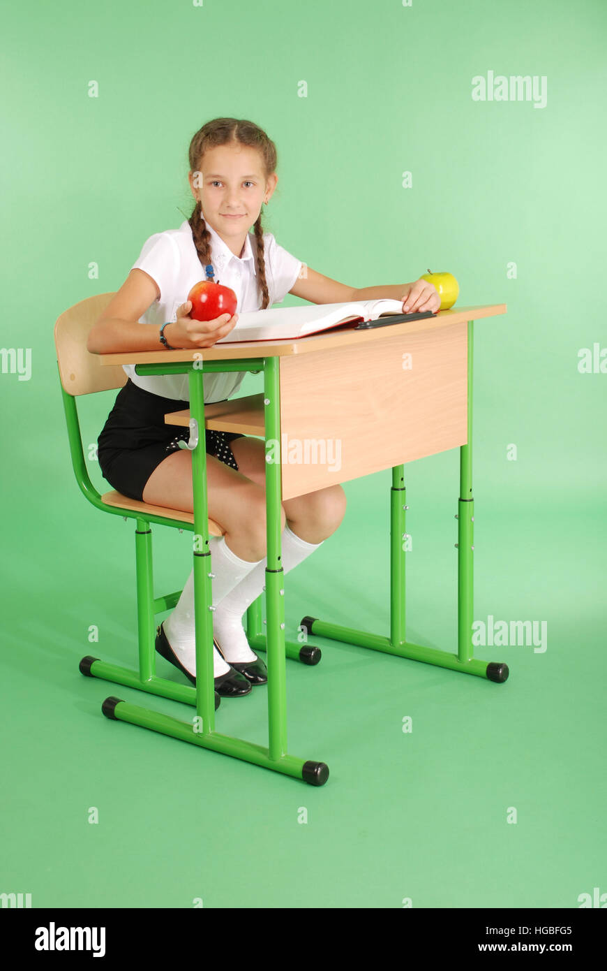 Girl in a school uniform sitting at a desk and reading a book isolated on greenGirl in a school uniform sitting at a desk and re Stock Photo
