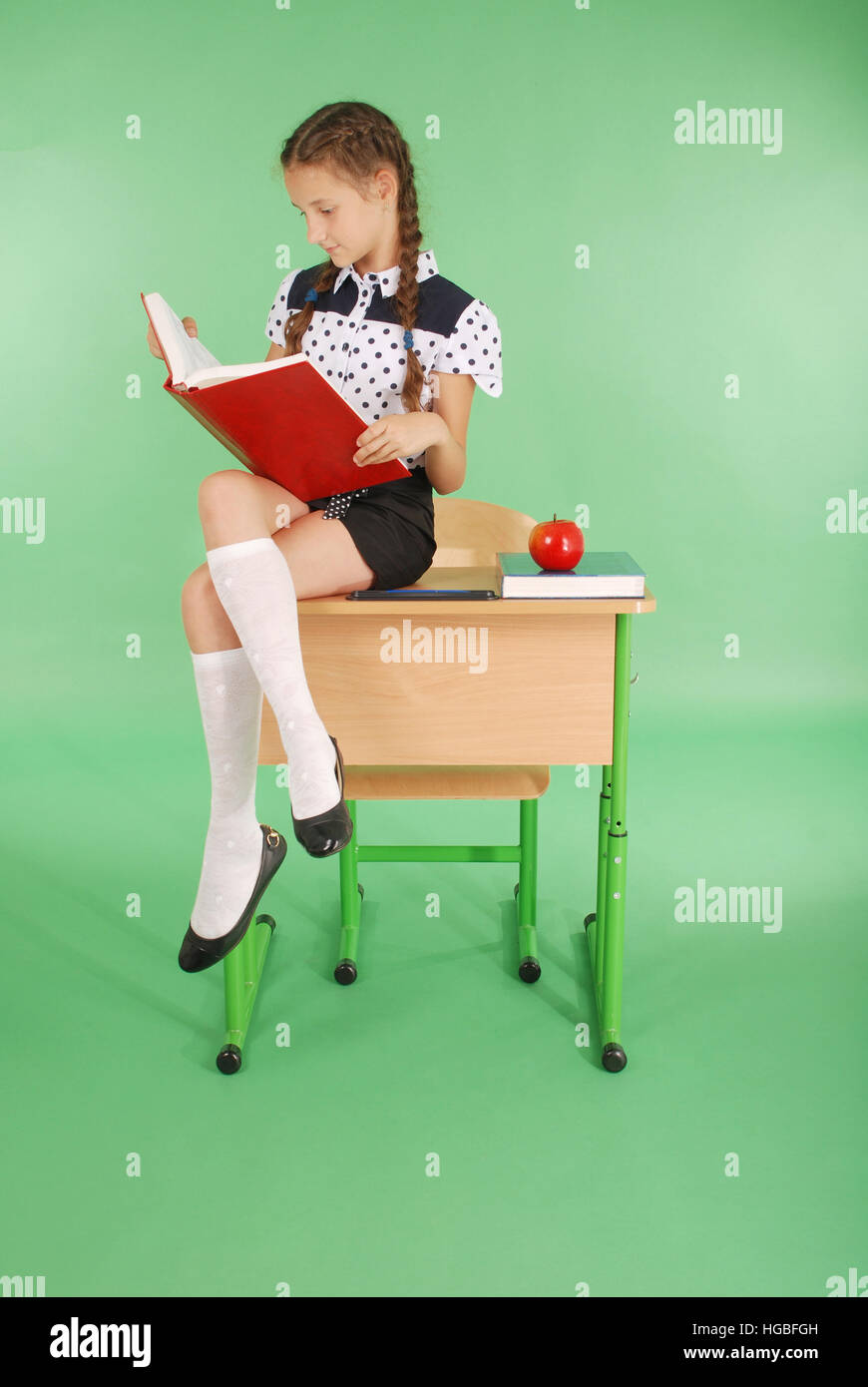 Girl in a school uniform sitting on desk and reading a book isolated on green Stock Photo