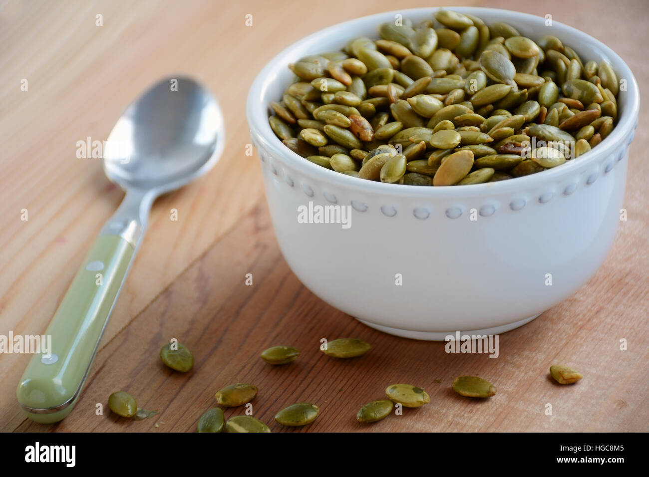 Roasted pumpkin seeds in white decorative bowl with green spoon on cedar background.  Horizontal format with shallow depth of field. Stock Photo