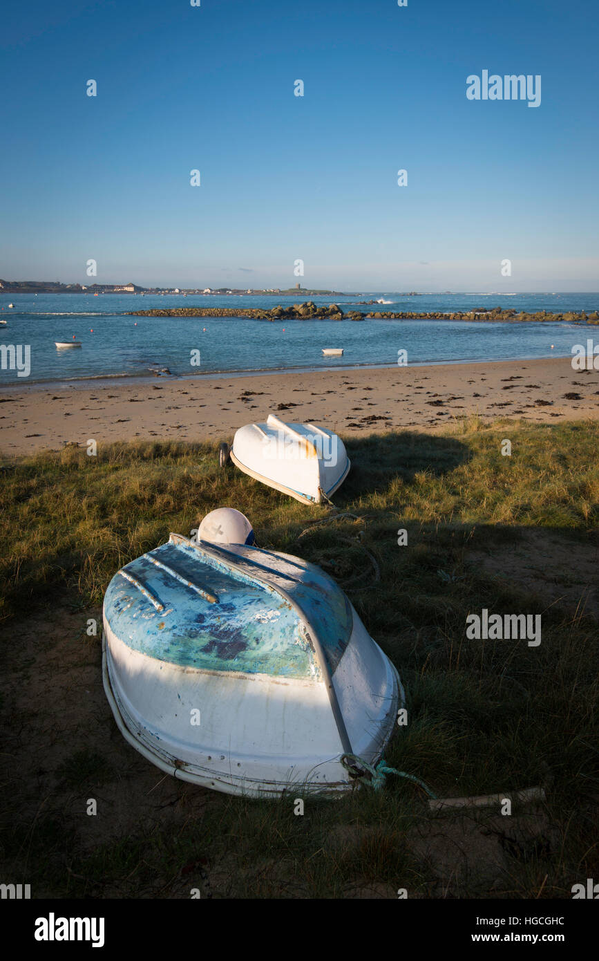 Small upturned boats on grass at top of beach, with rocks in the background and blue sky. Stock Photo