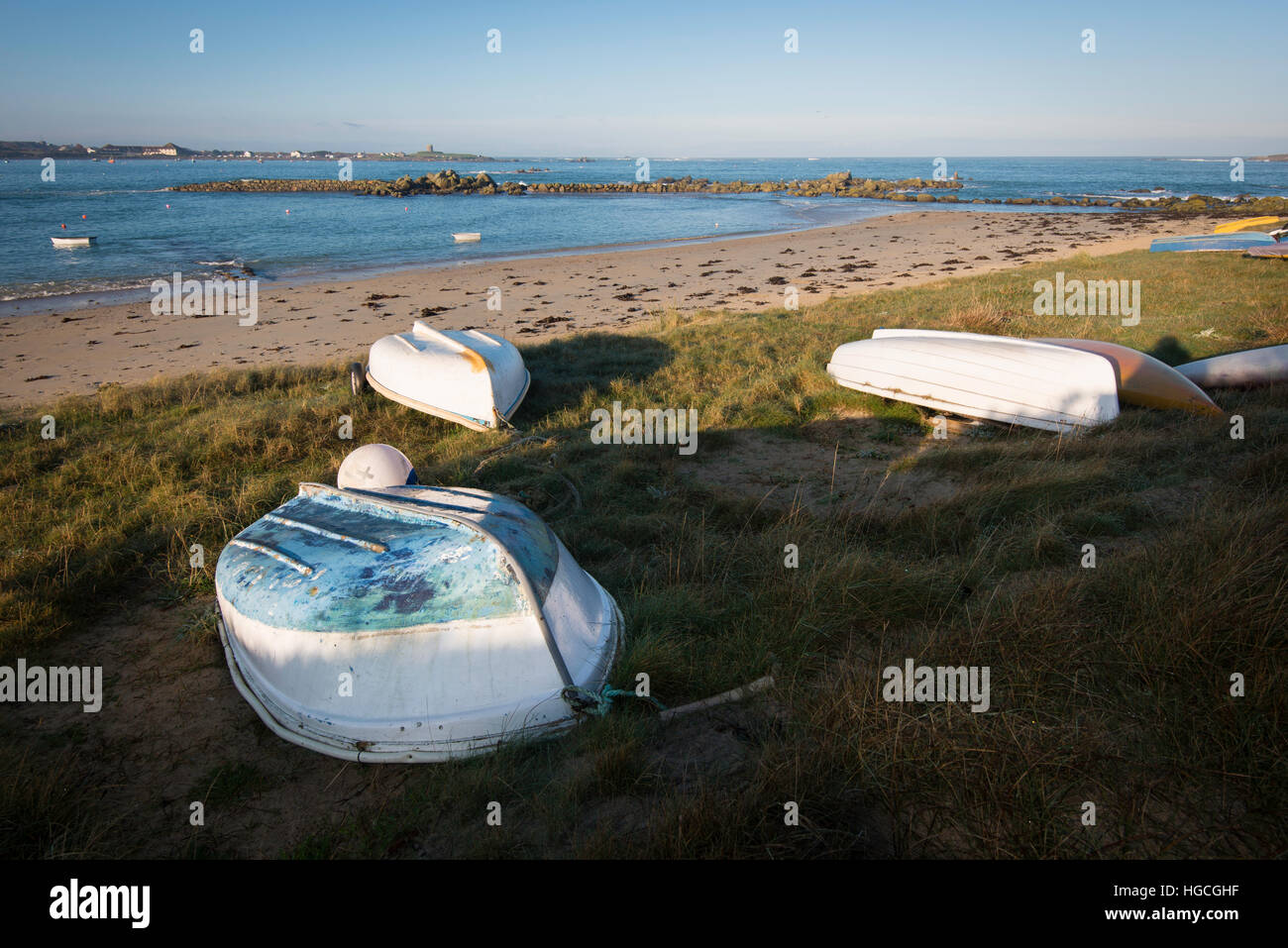 Small upturned boats on grass at top of beach, with rocks in the background and blue sky. Stock Photo