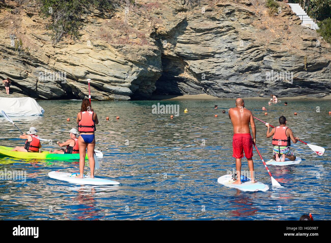 Holidaymakers on paddle boards with a small beach and cave to the rear, Bali, Crete, Greece, Europe. Stock Photo