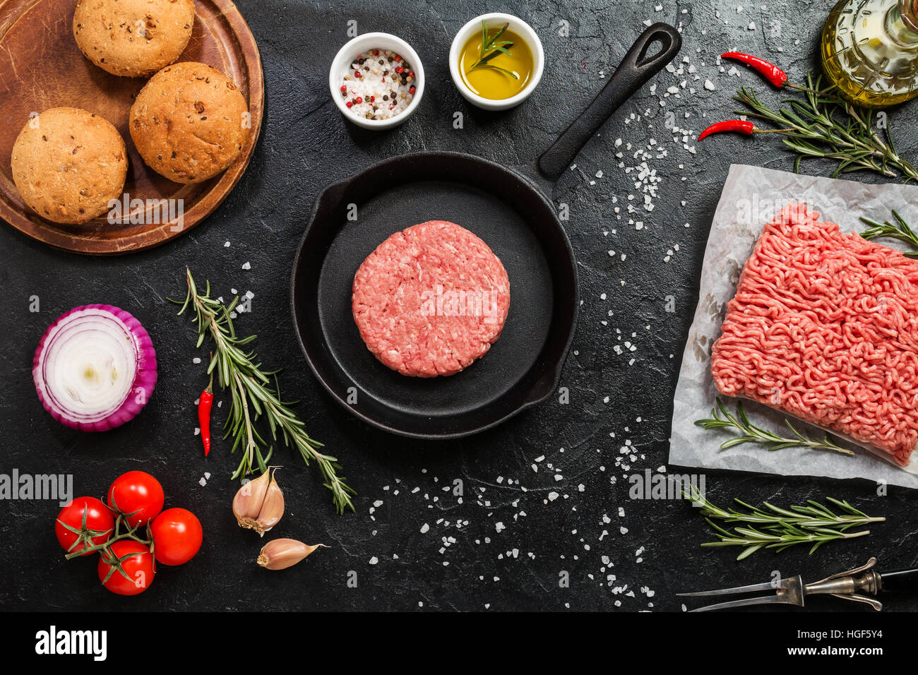 patty of minced meat on frying pan for burger with tomato, onion and seasonings on black background top view Stock Photo