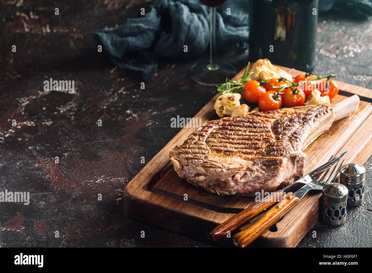 Succulent grilled tomahawk beef steak on the bone with red wine, seasonings, fresh rosemary and grilled vegetables on cutting board Stock Photo