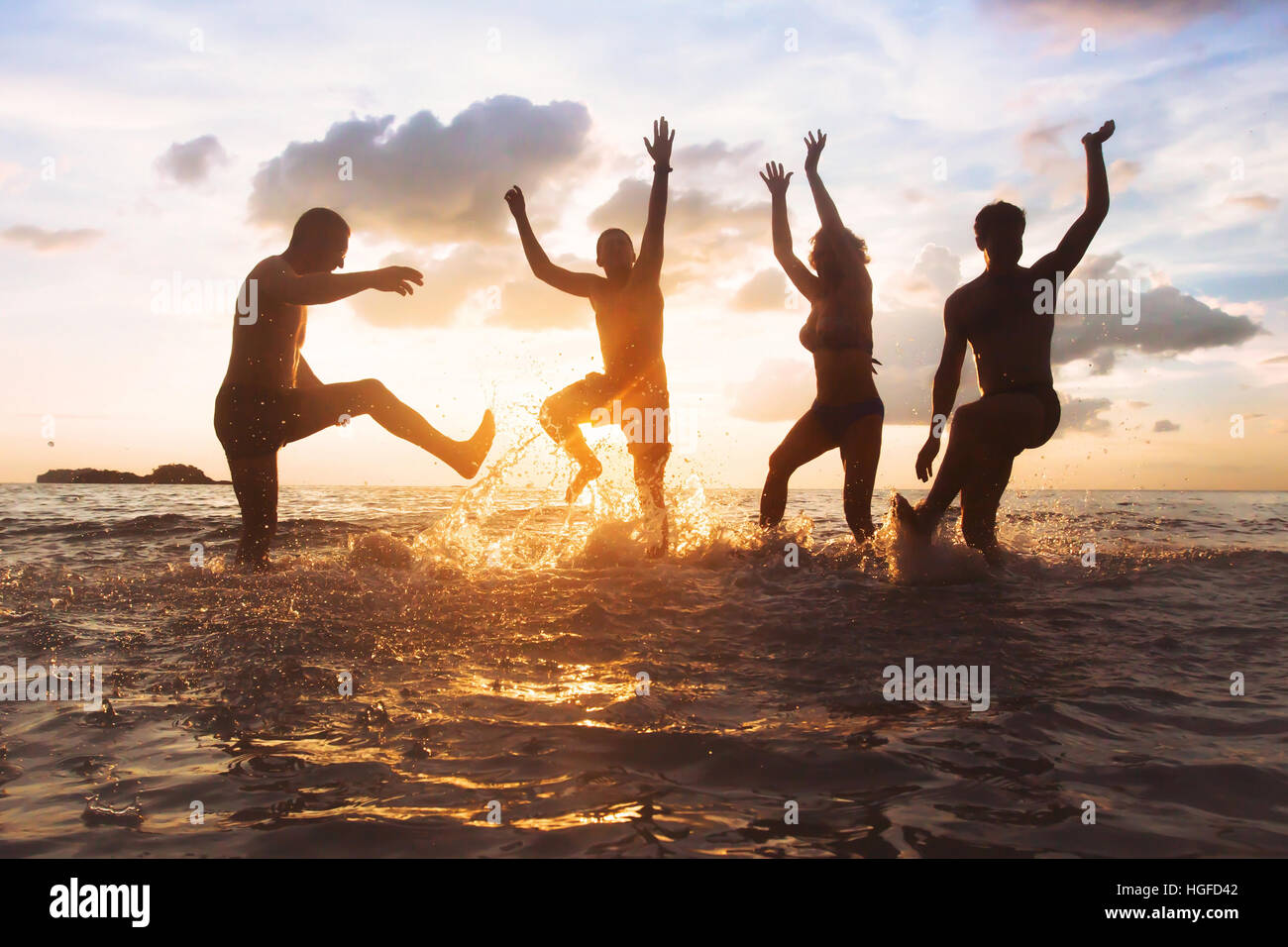 group of happy friends having fun together on the beach at sunset, jumping and dancing with water splash in the sea, silhouettes of people Stock Photo